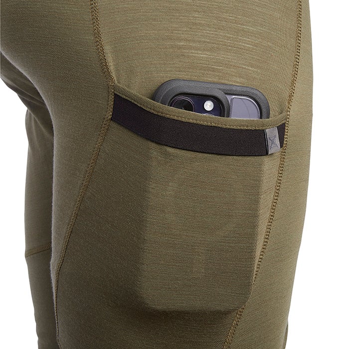 Close-up product shot of olive-green merino wool base layer leggings with a phone in a black elastic-banded pocket showing detailed stitching and a subtle texture