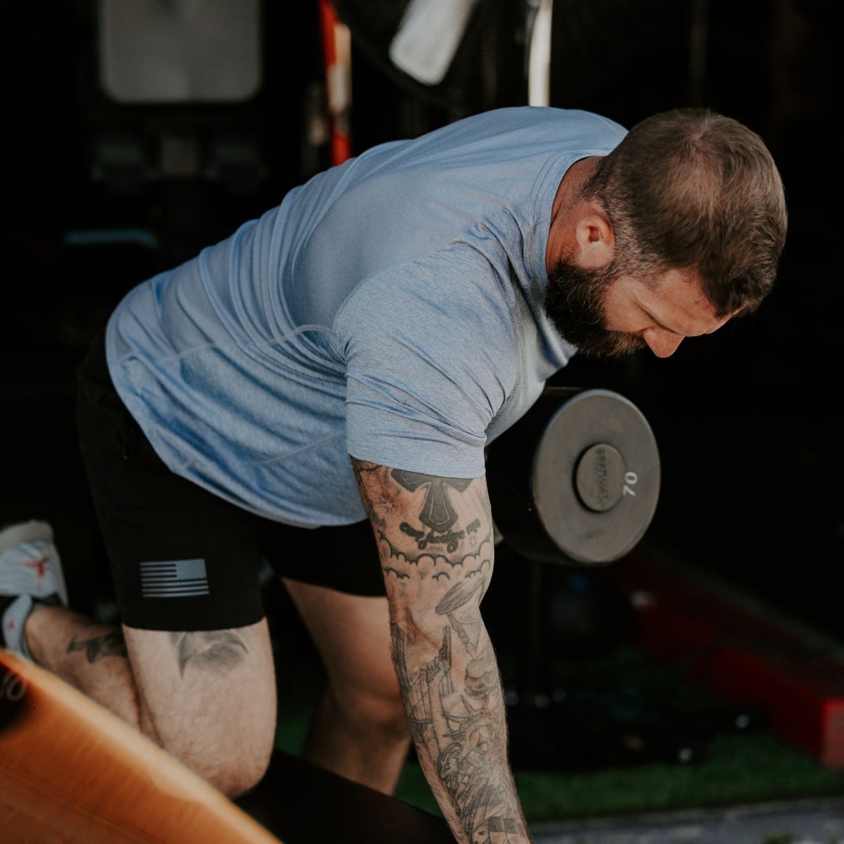 Photograph of a tattooed man performing a dumbbell row in a garage featuring a 20-pound weight, American flag detail on his shorts, and intricate sleeve tattoos