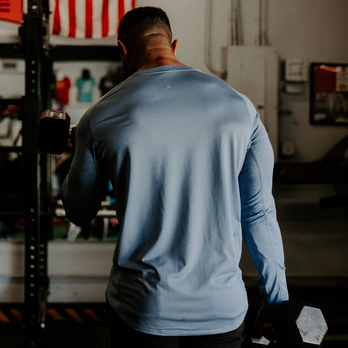 Photograph of a muscular man in a powder blue long-sleeve shirt lifting dumbbells in a gym with an American flag and weightlifting equipment visible in the background