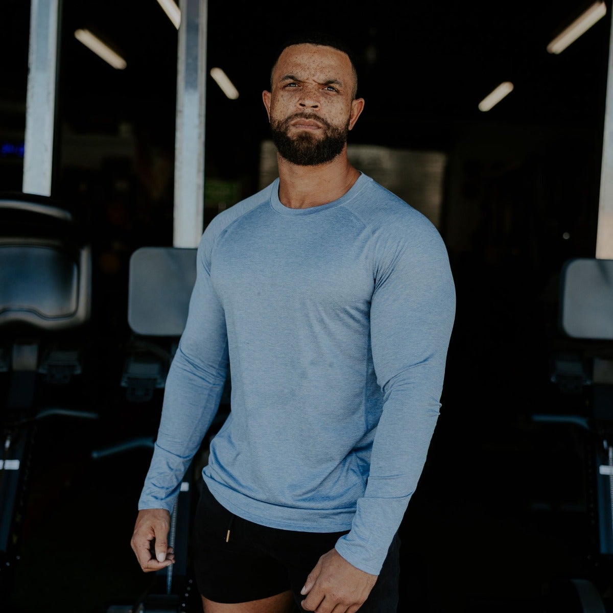 Photography Unknown Artist a muscular man in a light blue long-sleeve shirt and black shorts stands in a gym with dark-toned exercise equipment and bright overhead lighting showcasing his defined biceps and freckled complexion