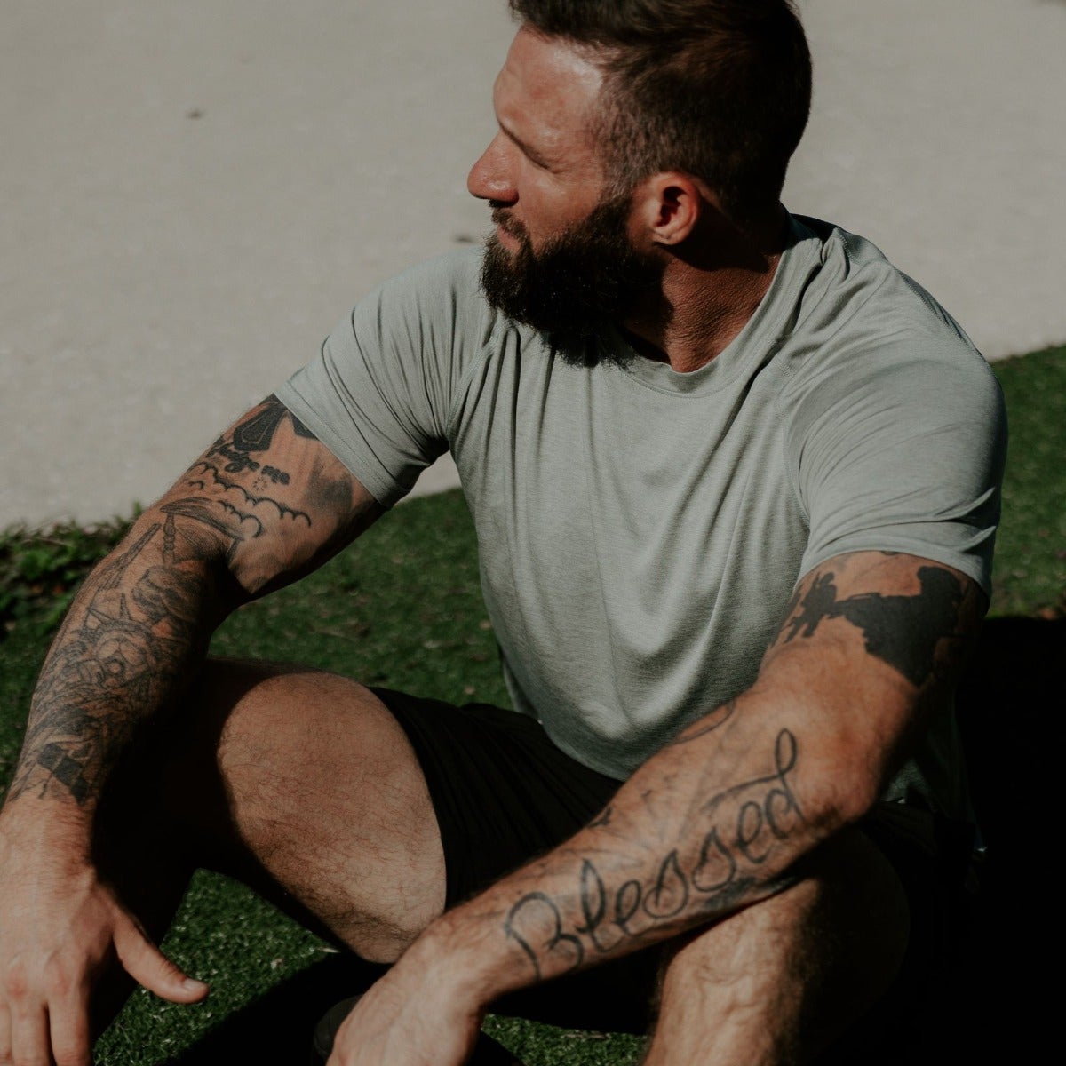 Photograph of a man with extensive arm tattoos including a world map and the word Blessed sitting outdoors on the grass wearing a sage green t-shirt and black shorts