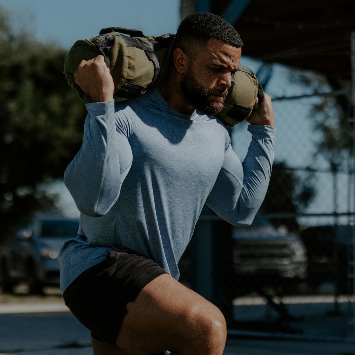 Photograph of a man in a blue shirt and black shorts carrying a heavy sandbag outdoors near a chain link fence and blurred cars showing his strong physique and determined expression