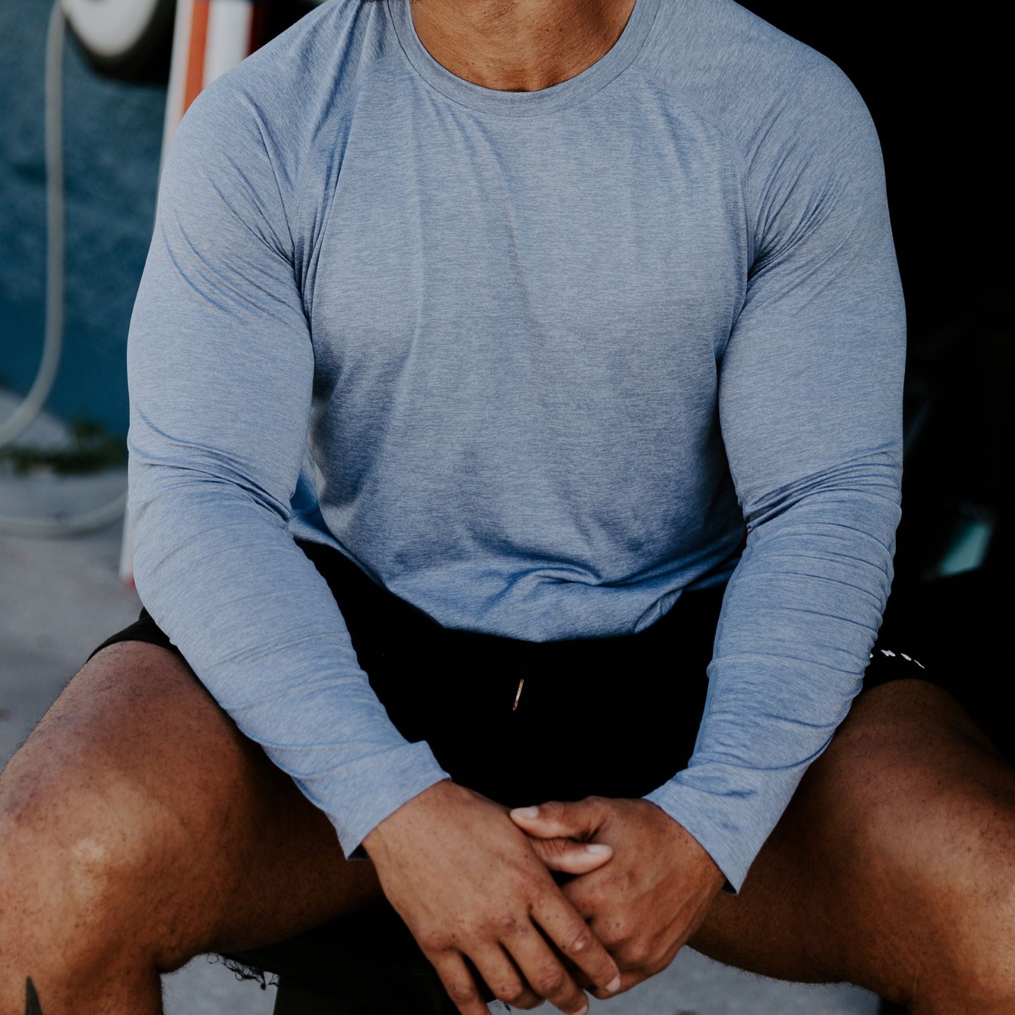Photograph of a muscular man wearing a heather blue long-sleeve shirt and black shorts seated outdoors with subtly textured fabric and sunlit skin tones showing a hint of dark-haired legs