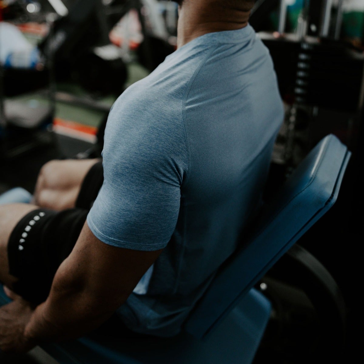 Photograph of a muscular man in a blue heather athletic shirt seated on a leg press machine in a gym with dark-toned workout equipment and a subtly textured fabric