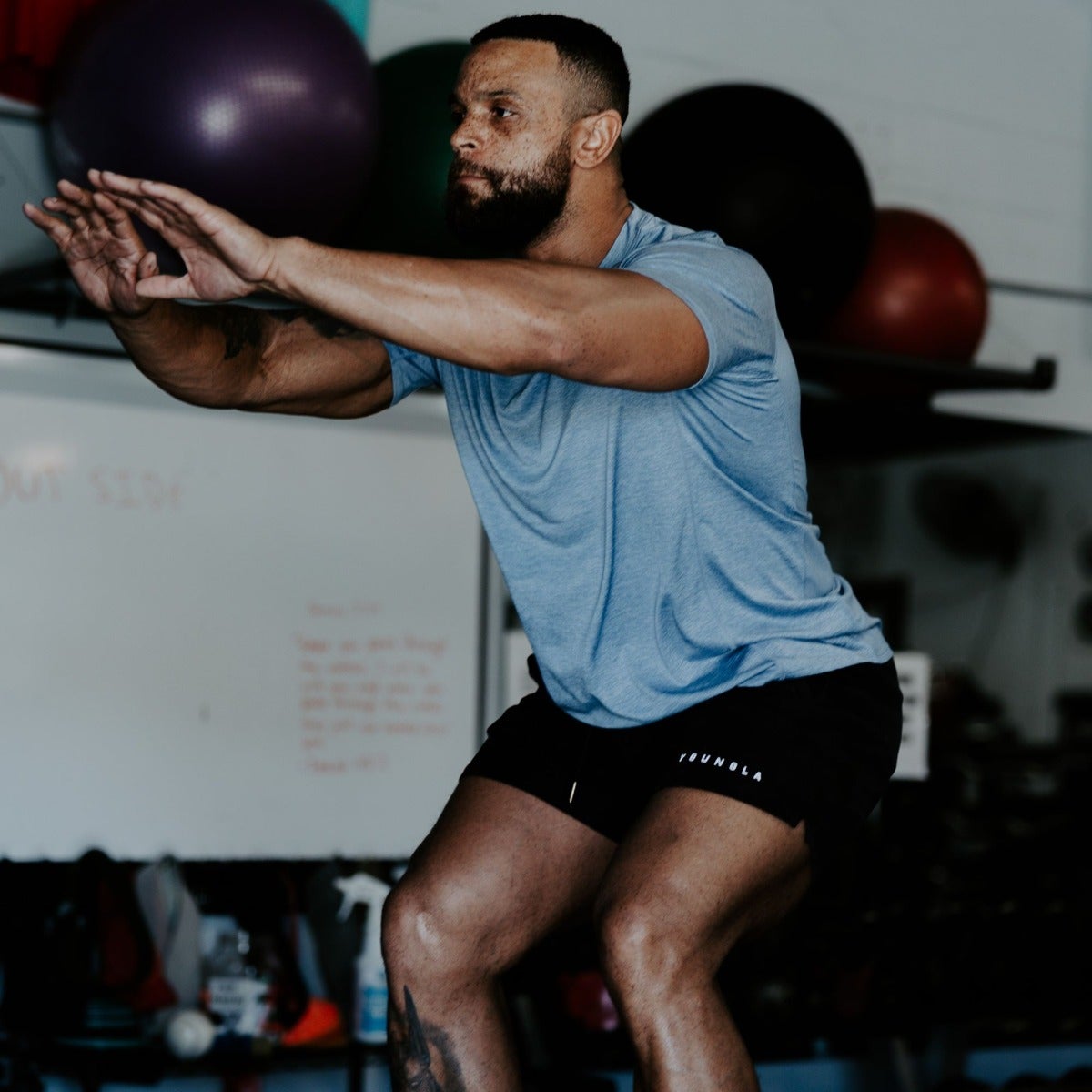 Photograph of a man performing a squat exercise in a gym with purple and red exercise balls a whiteboard and a partially visible tattoo on his leg