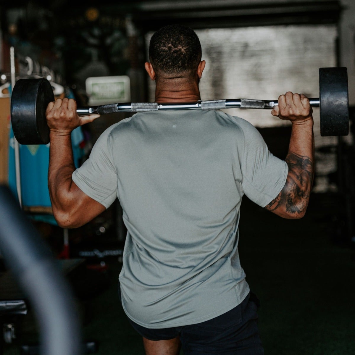 Photograph of a man in a sage green shirt performing a barbell back lift in a home gym featuring a dark gray wall teal clothing and a detailed arm tattoo