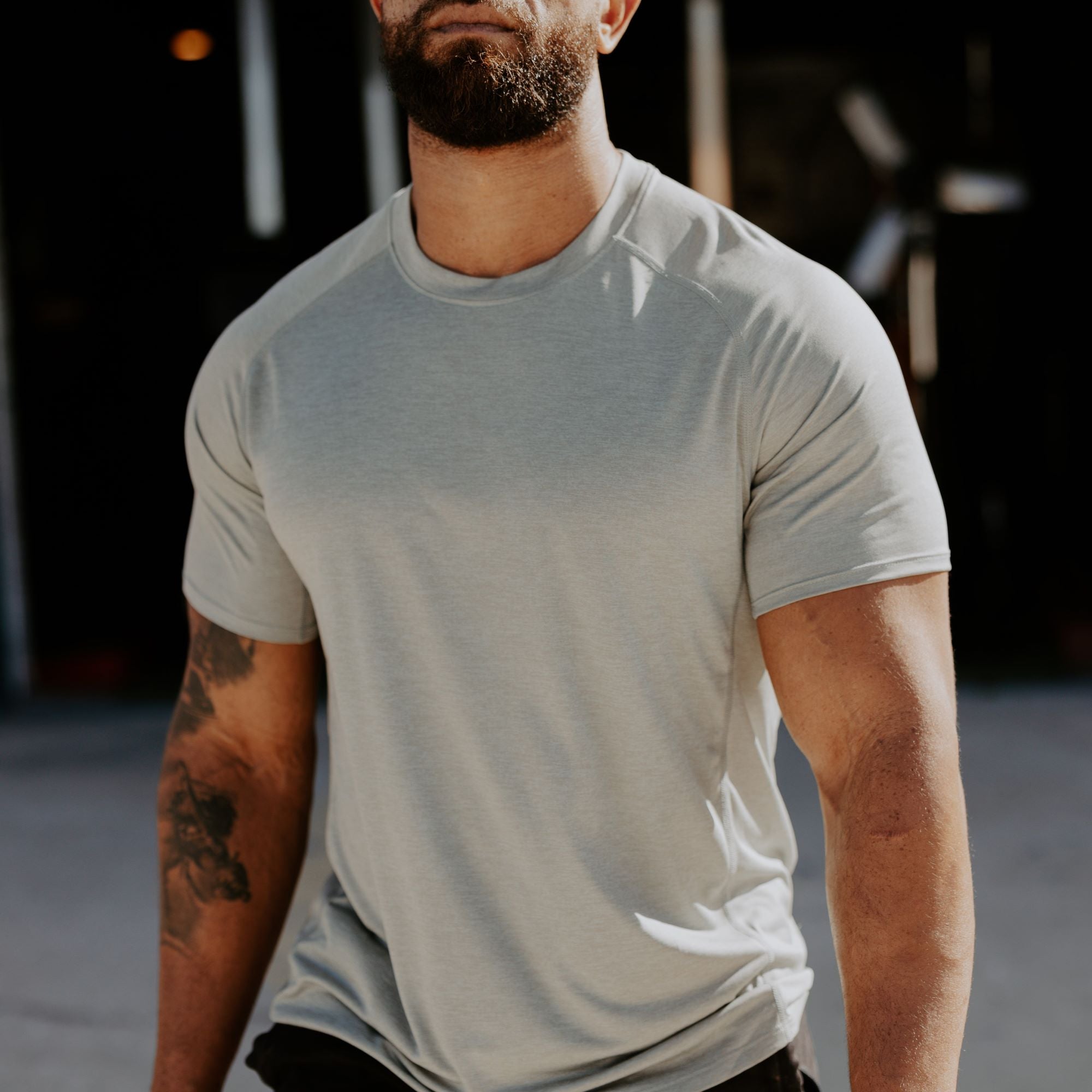 Photograph of a man wearing a light grey, athletic fit t-shirt outdoors, showcasing his muscular physique, a detailed arm tattoo, and a subtle scar, against a blurred background of industrial elements