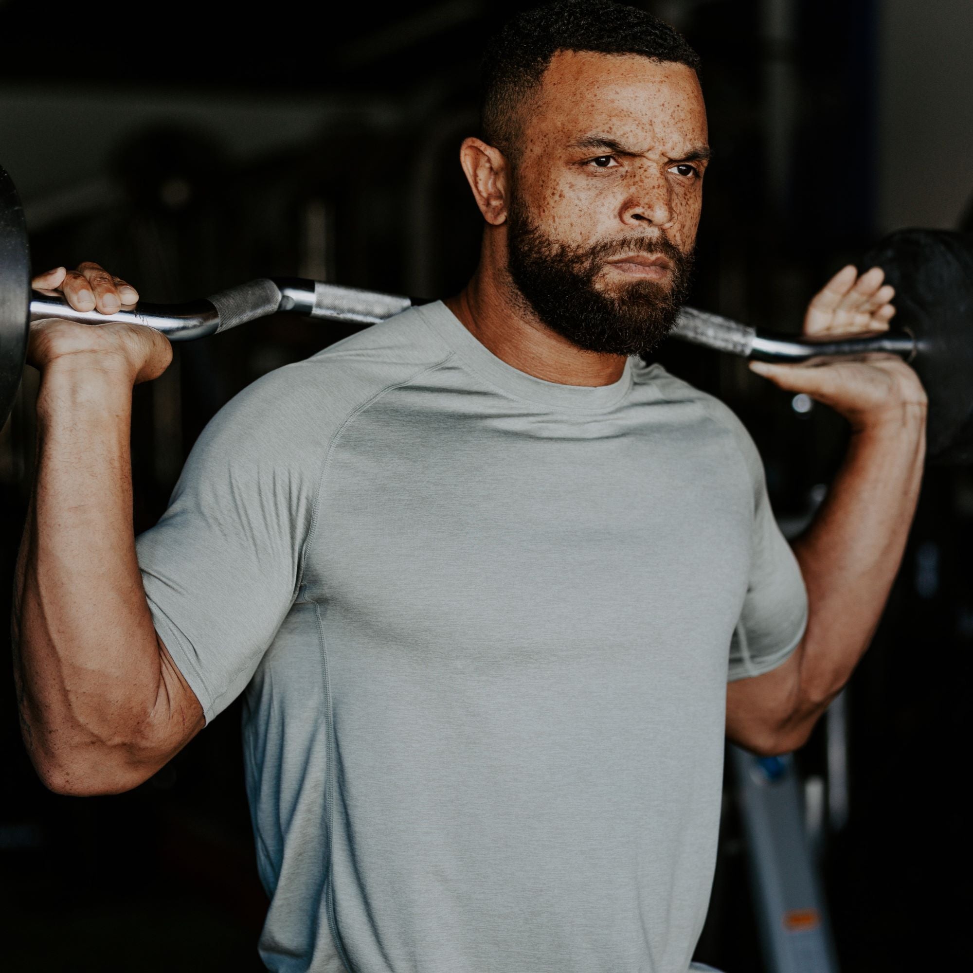 Photograph of a man in a sage green shirt performing a barbell back squat in a dimly lit gym, showcasing his defined muscles, freckled skin, and a focused expression