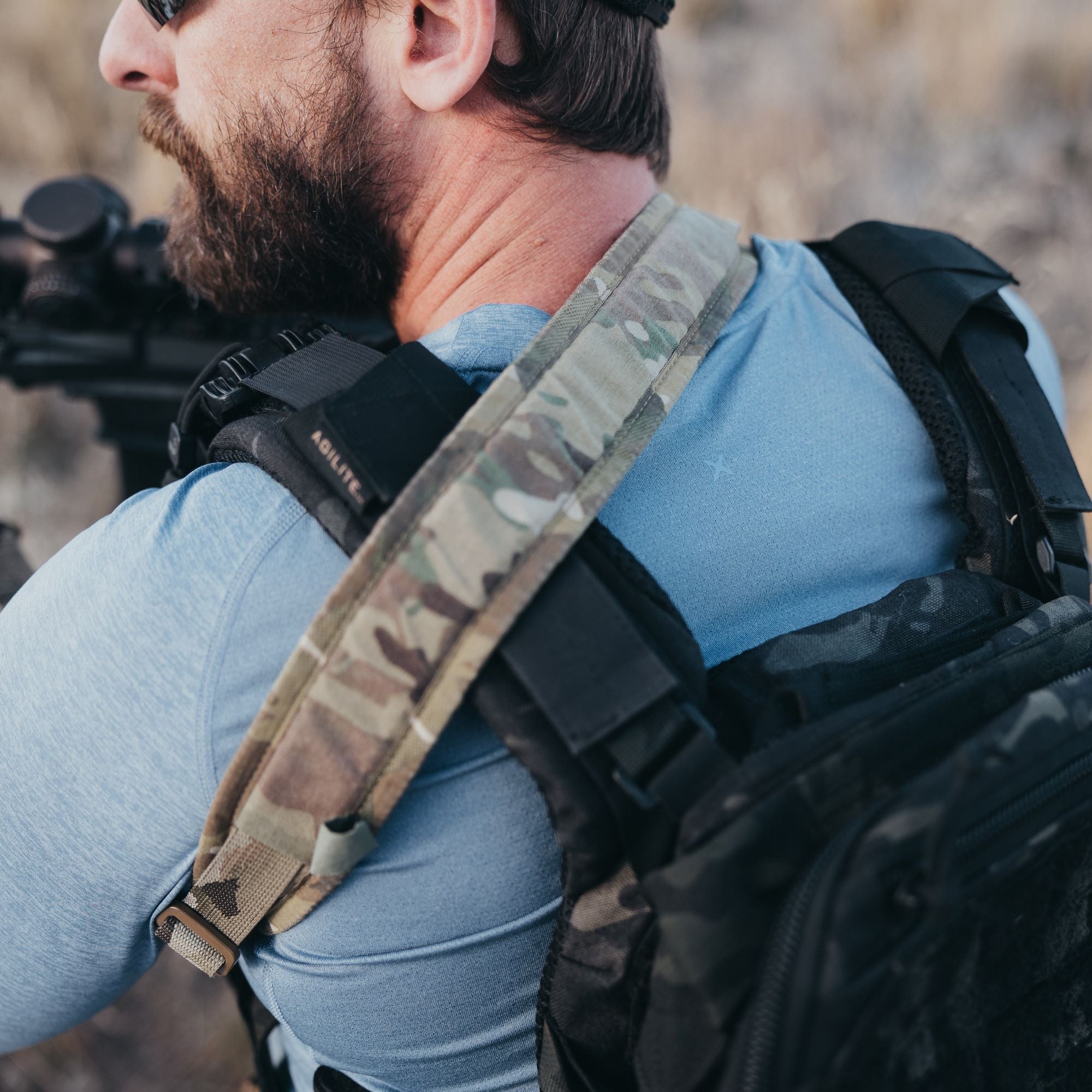 Photography shows a man outdoors wearing a blue shirt and a camouflage backpack with a brown buckle and black Agilite tag, carrying a rifle with a scope