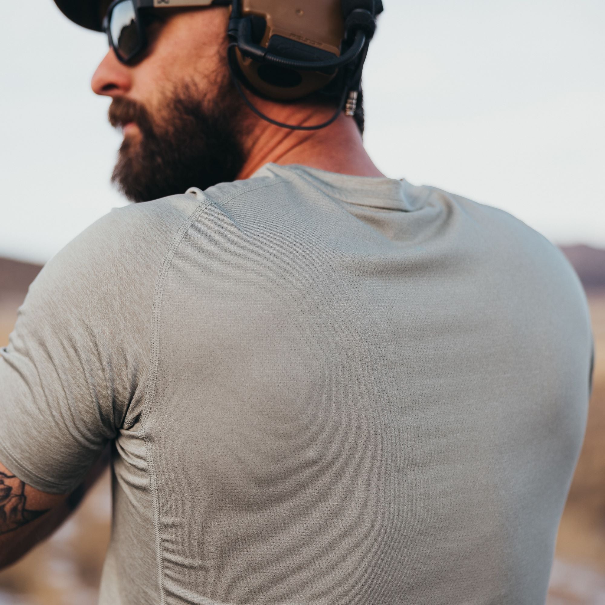 A photograph showing the back of a man wearing a light gray performance shirt with unique stitching details and a subtle texture in a desert landscape with mountains in the distance