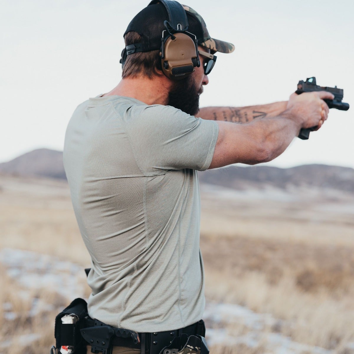 Photograph of a man in a sage green shirt and camouflage hat firing a handgun in a desert landscape with distant mountains and a textured fabric shirt