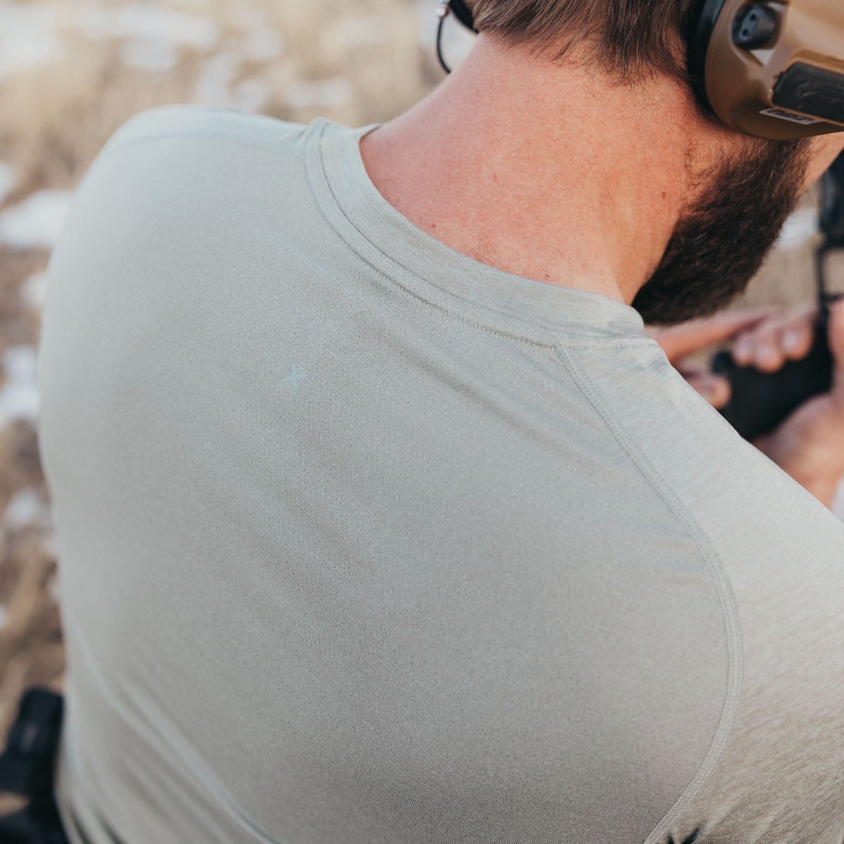 Photograph of a man's back wearing a light sage-green performance shirt with a small, faint mark near the shoulder, showing his neck and part of his beard, and a tan-colored background