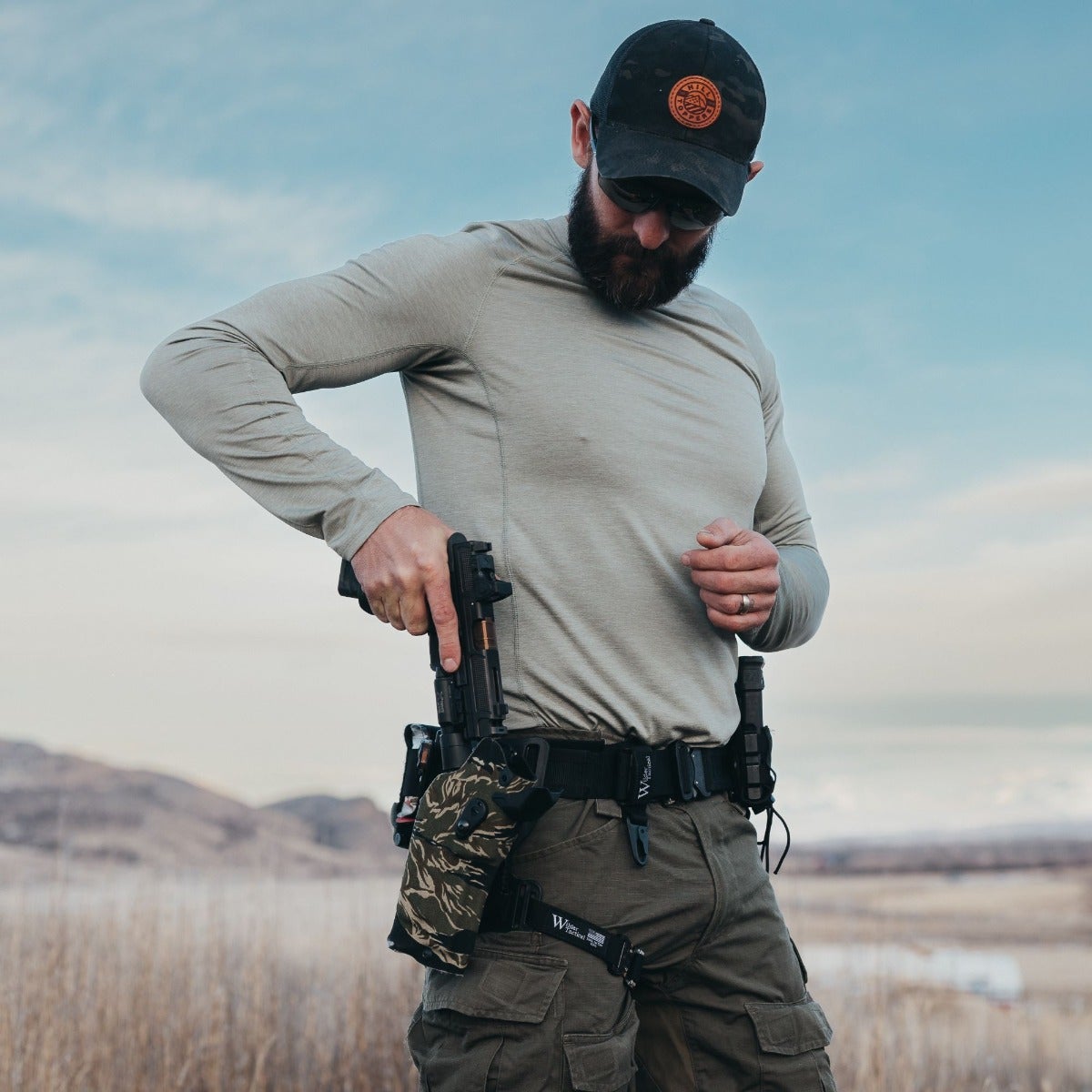 A photograph of a man with a beard, wearing a grey long-sleeve shirt and a black cap, holsters a handgun against a backdrop of high-desert hills and a pale blue sky featuring a camouflage-patterned holster, a black tactical belt, and a brown leather patch on his hat