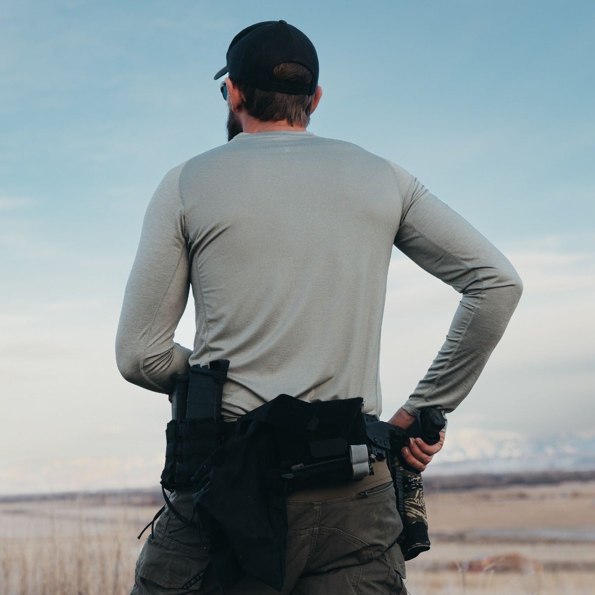 Photograph of a man in a gray long-sleeve shirt and tactical gear overlooking a vast landscape with distant mountains under a pale blue sky featuring a black baseball cap, olive green pants and a dark handgun in a holster.