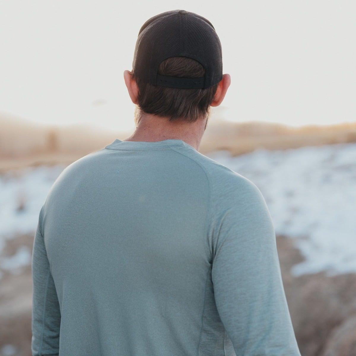 Photograph of a man wearing a sage green long-sleeved shirt and black baseball cap overlooking a snowy landscape with muted golden hills in the background