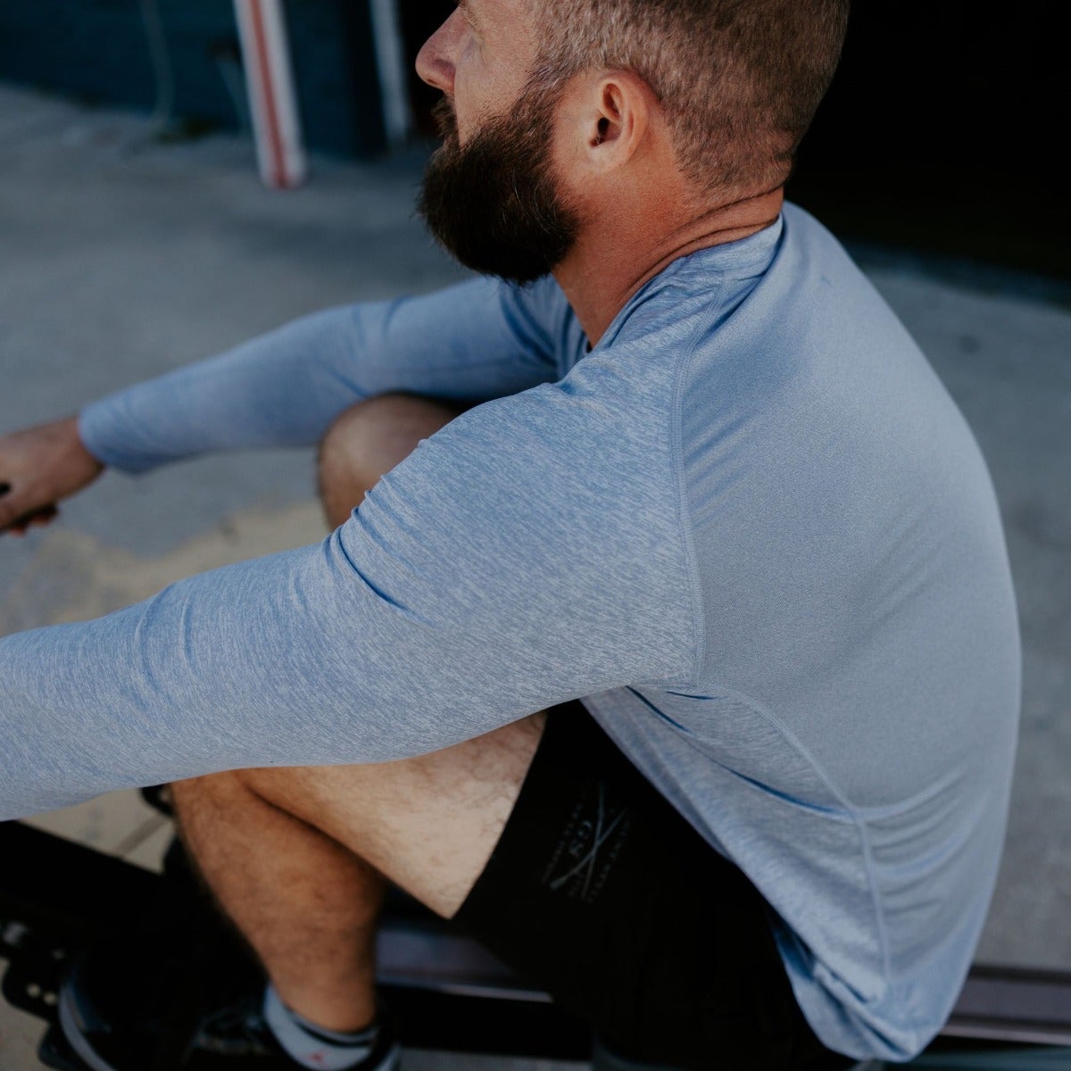 Photography of a man wearing a heather blue long-sleeve shirt and black shorts while sitting on a rowing machine outdoors showcasing the shirt's texture and comfortable fit.