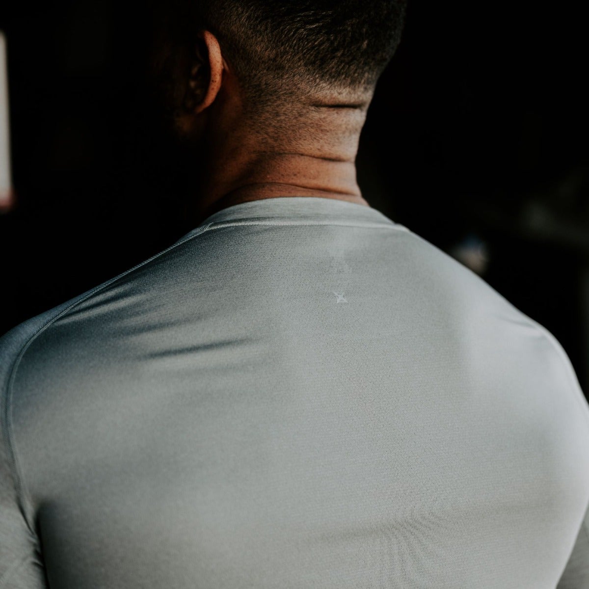 Photograph of a man's back wearing a pale grey shirt with a small, faint marking near the center featuring subtle shadows and a textured fabric