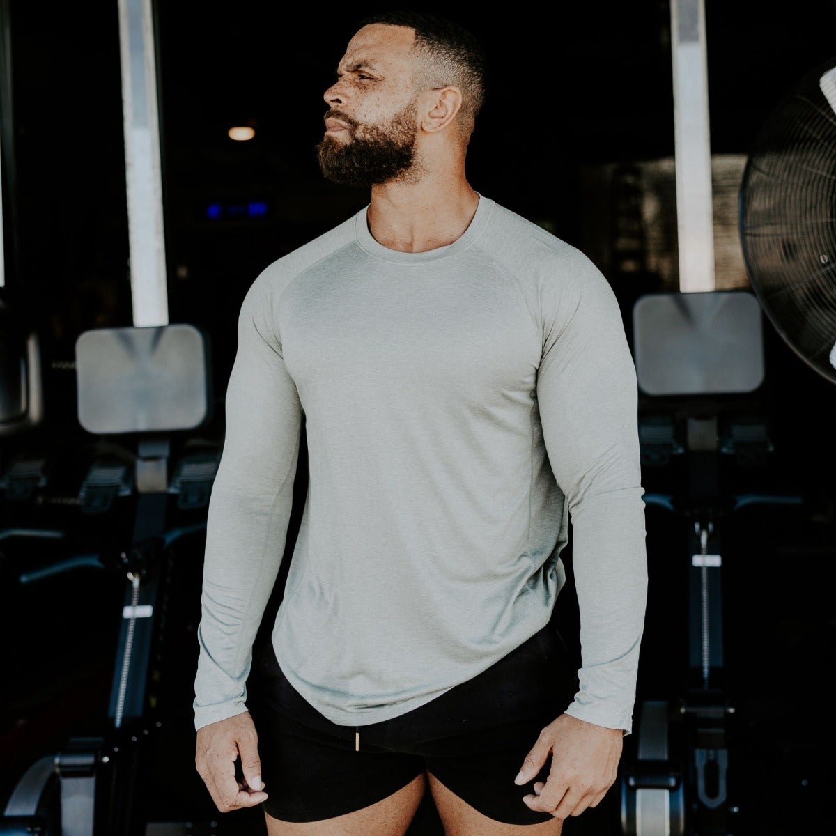 Photography Unknown Artist a muscular man in a sage green long-sleeve shirt and black shorts stands in a gym with rowing machines and a large industrial fan showcasing his defined biceps and beard