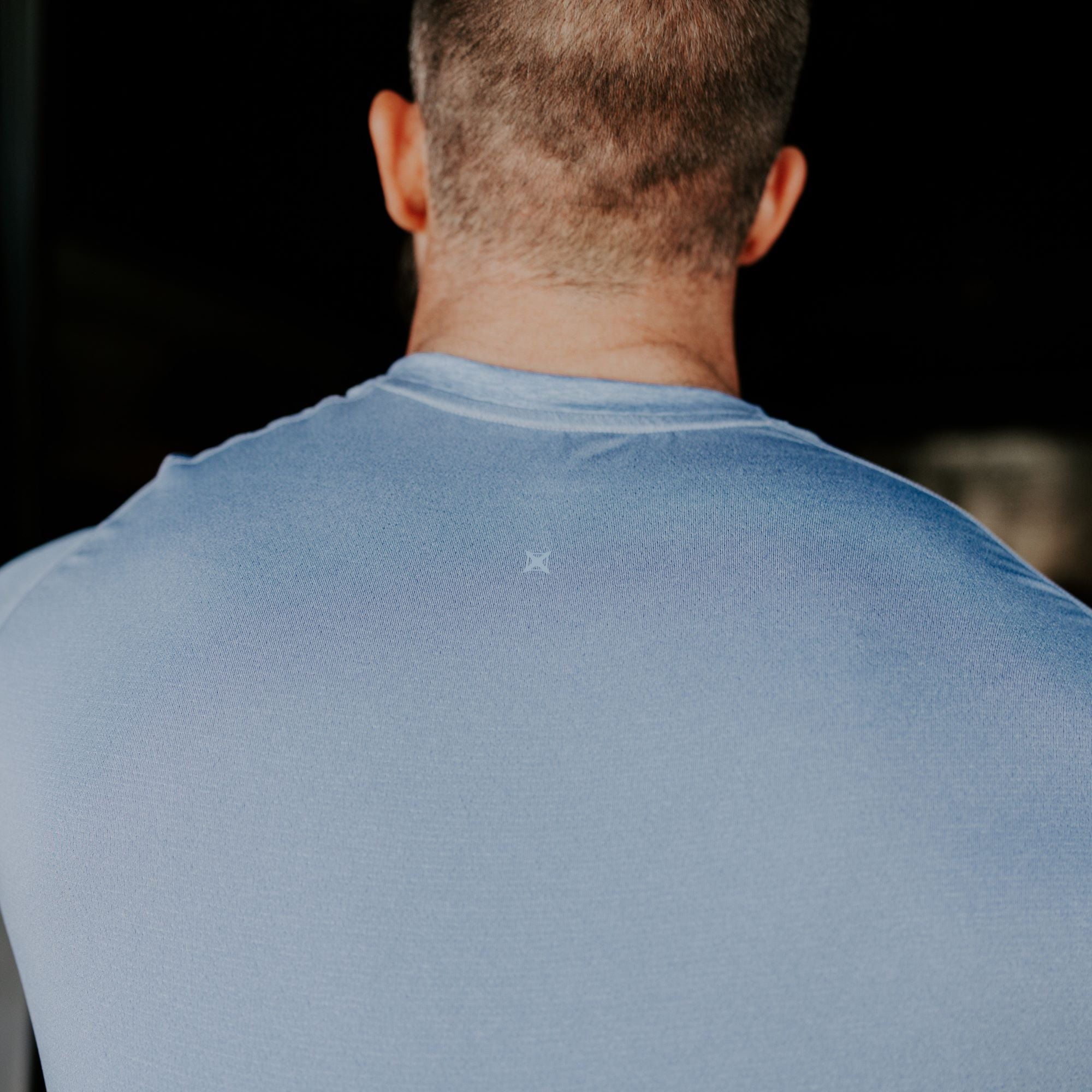 Photograph of a man's back wearing a pale blue shirt with a small, barely visible logo, showcasing his short brown hair and the texture of the fabric
