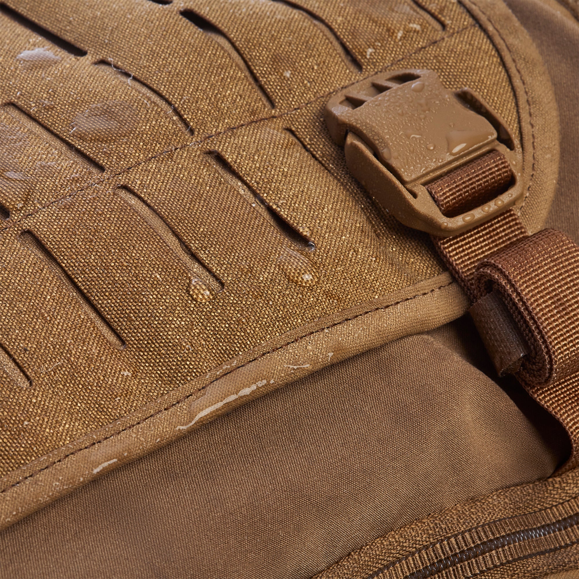 Close-up of coyote brown waterproof fabric with water droplets beading, showing textured MOLLE webbing slots and a matching plastic buckle