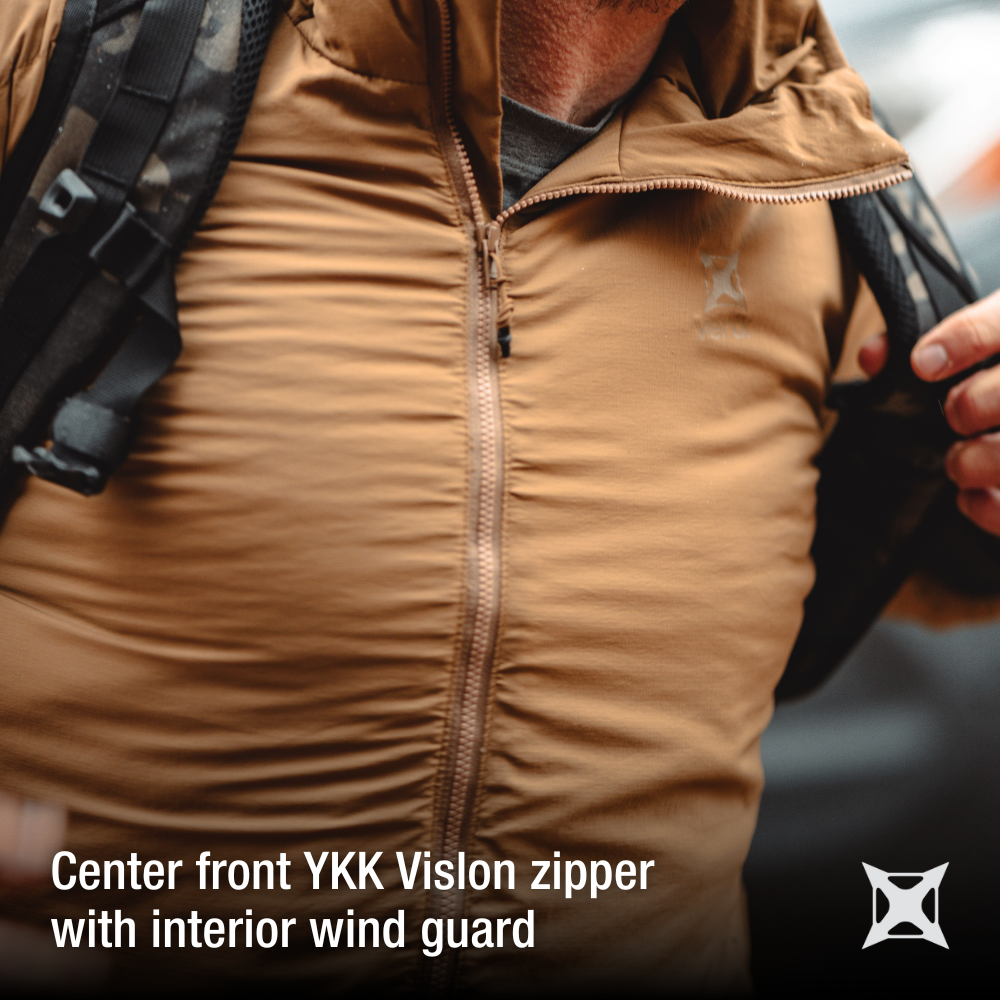 Close-up photograph showing a tan jacket with a YKK Vislon zipper and interior wind guard detail a person wearing it outdoors with a camouflage backpack the fabric has a subtle texture and the zipper is a warm copper tone