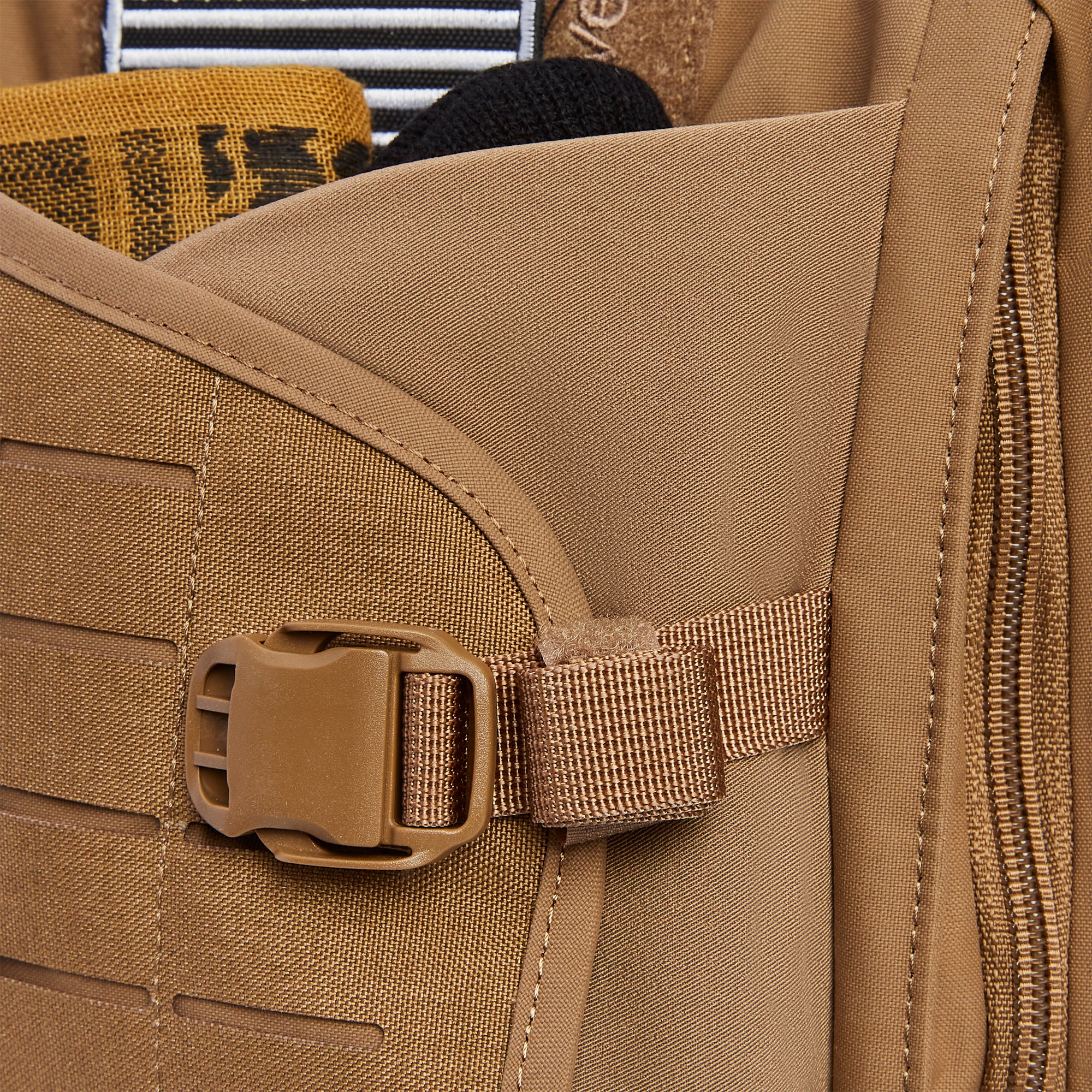Close-up of a coyote brown tactical backpack showcasing a plastic quick-release buckle, MOLLE webbing, a prominent side zipper, and glimpses of a yellow woven fabric with black text and a black and white striped patch peeking from an opening