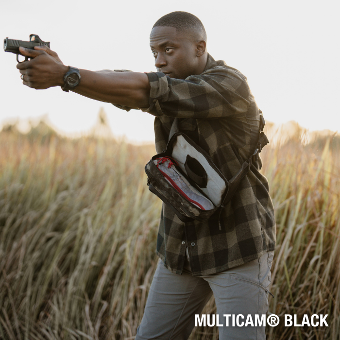 Photograph of a man in a field aiming a handgun wearing a Multicam Black tactical chest rig with a gray watch and olive green plaid shirt