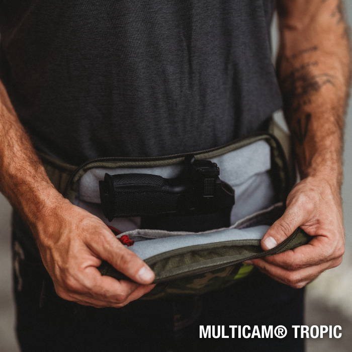 Photograph showing a man holding open a Multicam Tropic fanny pack revealing a black handgun inside a gray padded compartment with red stitching and olive green exterior
