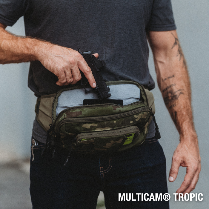 Photograph showing a person placing a black handgun into a Multicam Tropic camouflage fanny pack with a gray interior and lime green accents against a gray wall