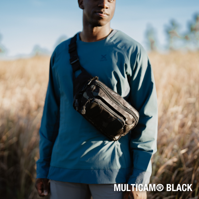 Photograph of a person wearing a Multicam Black crossbody bag in a field of tall grasses under a clear blue sky
