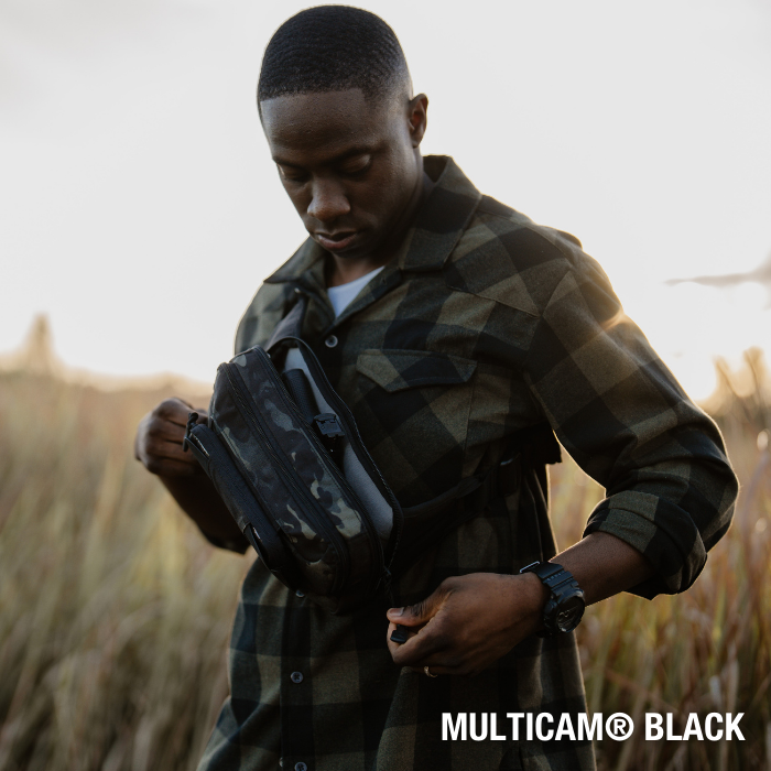 A photograph shows a man in a green and black plaid shirt adjusting a Multicam Black crossbody bag in a field with tall grasses at sunset