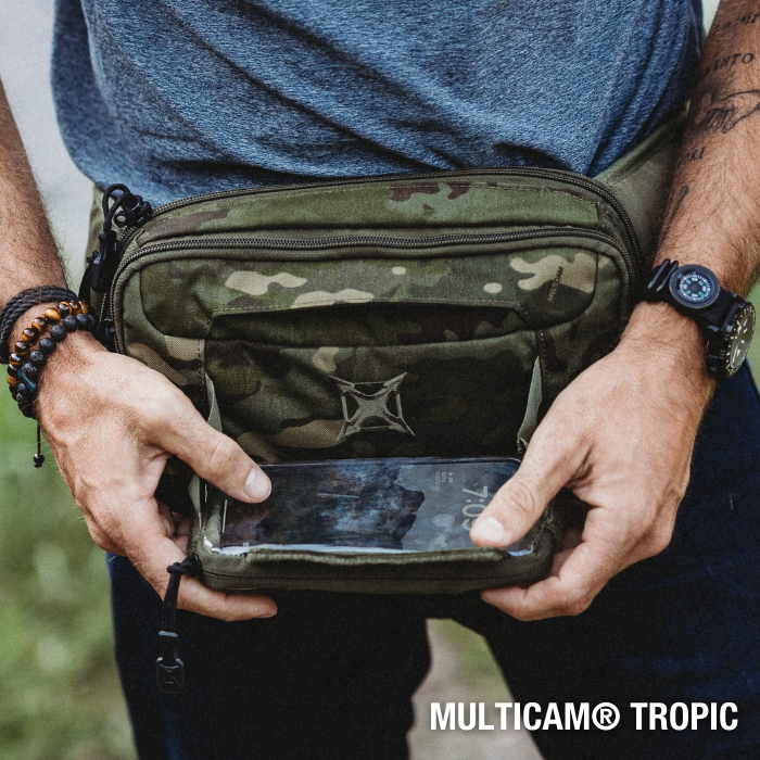Photograph showing a person wearing a Multicam Tropic fanny pack featuring a visible smartphone in a clear pocket and a distinctive cross emblem with brown beaded bracelets and a compass watch