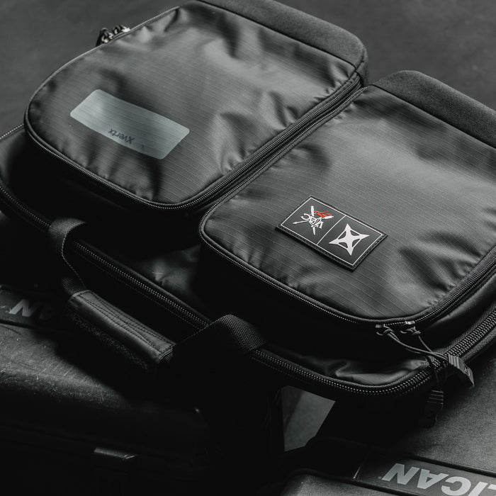 Product photography of a black XTRX double-compartment drum hardware bag featuring a gray XTRX label, embroidered logo patch, and reinforced zippers on a dark surface