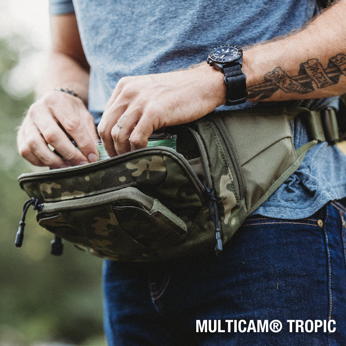 Photograph of a person wearing a Multicam Tropic camouflage fanny pack outdoors showing a black watch a tattooed arm and detailed zipper pulls