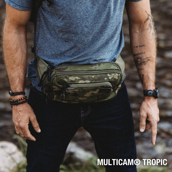 Photograph of a man wearing a Multicam Tropic fanny pack outdoors near rocks and water featuring a camouflage pattern, multiple zippered compartments, and dark denim jeans