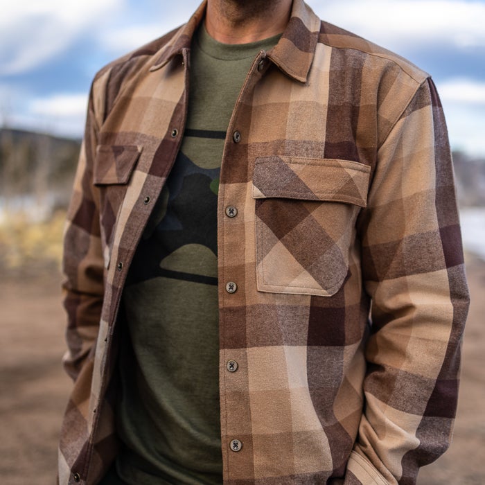 Photograph of a man wearing a brown and tan plaid flannel shirt outdoors with muted earth tones and subtle texture against a blurred background of hills