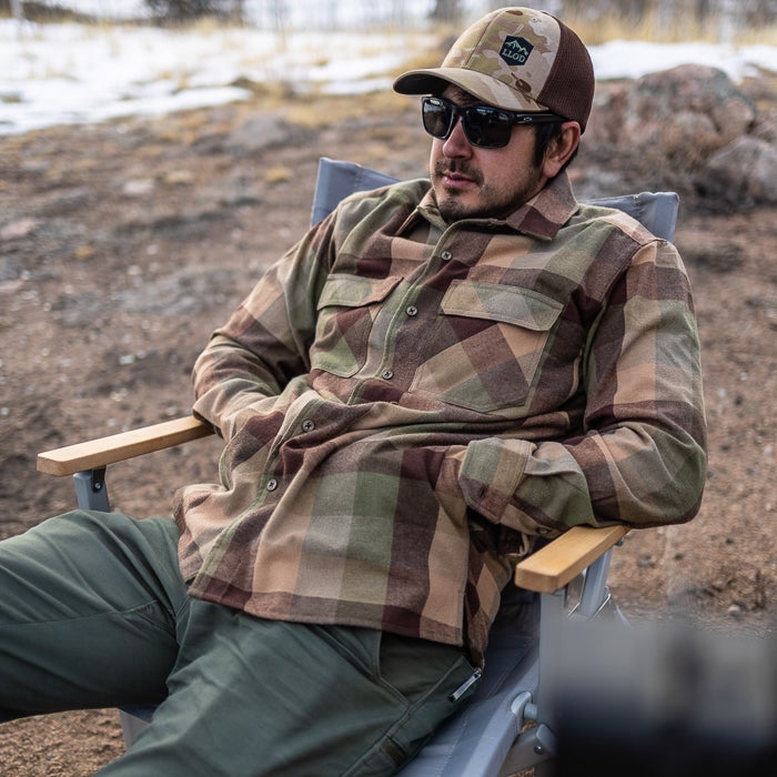 Photograph man wearing tan brown and green plaid shirt and sunglasses sitting in a wooden camping chair outdoors against a background of snowy hills
