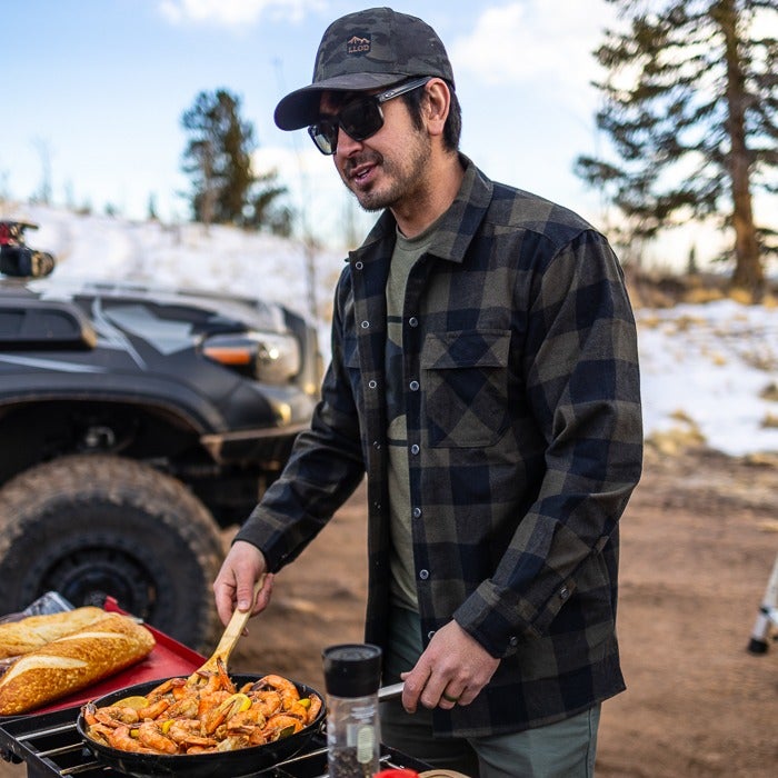 Photograph man in a dark plaid shirt cooking shrimp outdoors near a truck and snowy pines with a rustic wooden cooking setup and bright yellow lemon slices