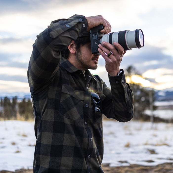 Photograph of a man in a dark green and black plaid shirt using a Canon camera with a large lens to capture a snowy landscape with distant mountains and a setting sun