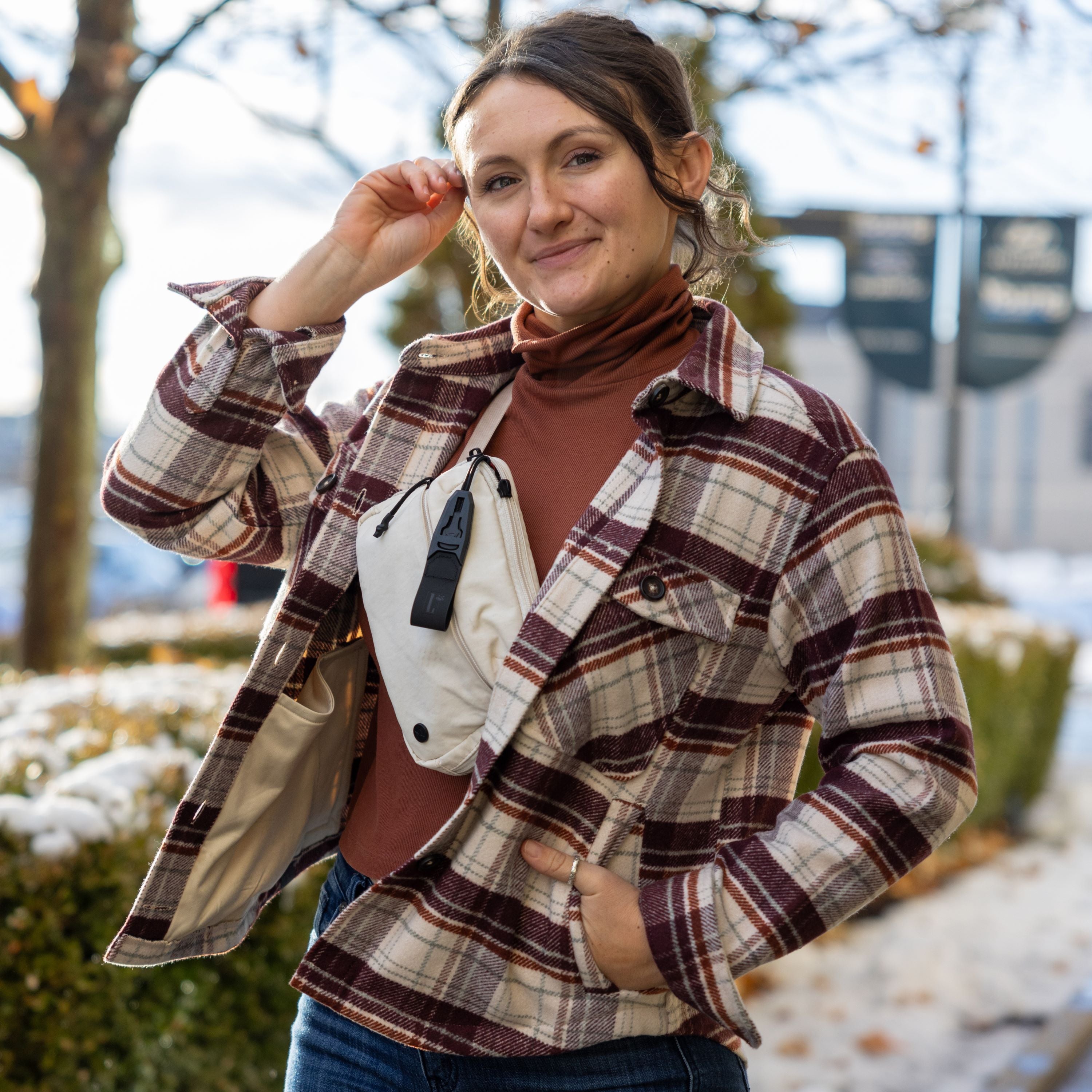 Photograph of a woman wearing a burgundy and cream plaid shirt, a terracotta turtleneck, and a beige fanny pack, standing outdoors near blurred buildings and trees with a hint of snow.