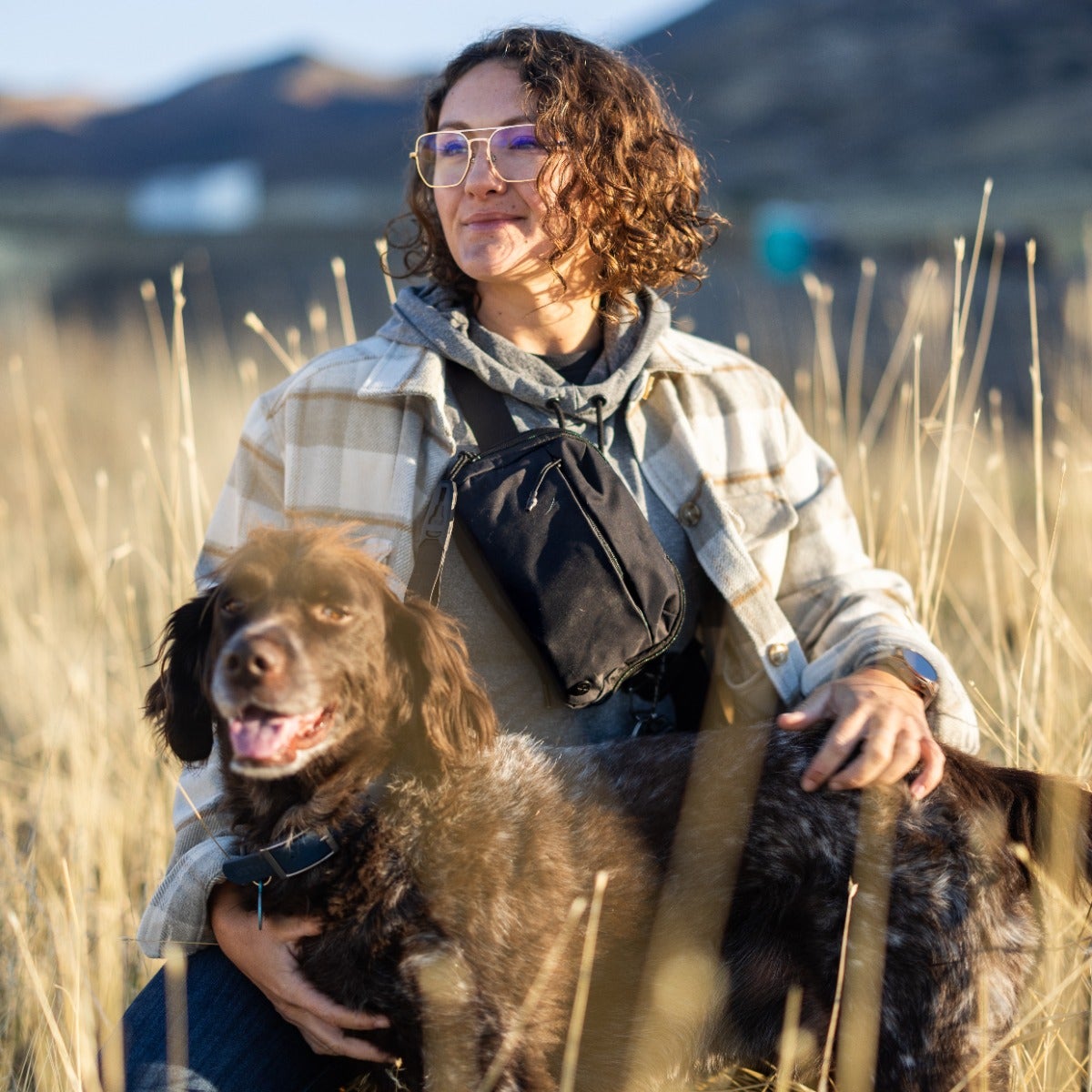 Photograph of a woman with a brown and white dog in a field with golden grasses and distant mountains showing blue skies and a hint of highway