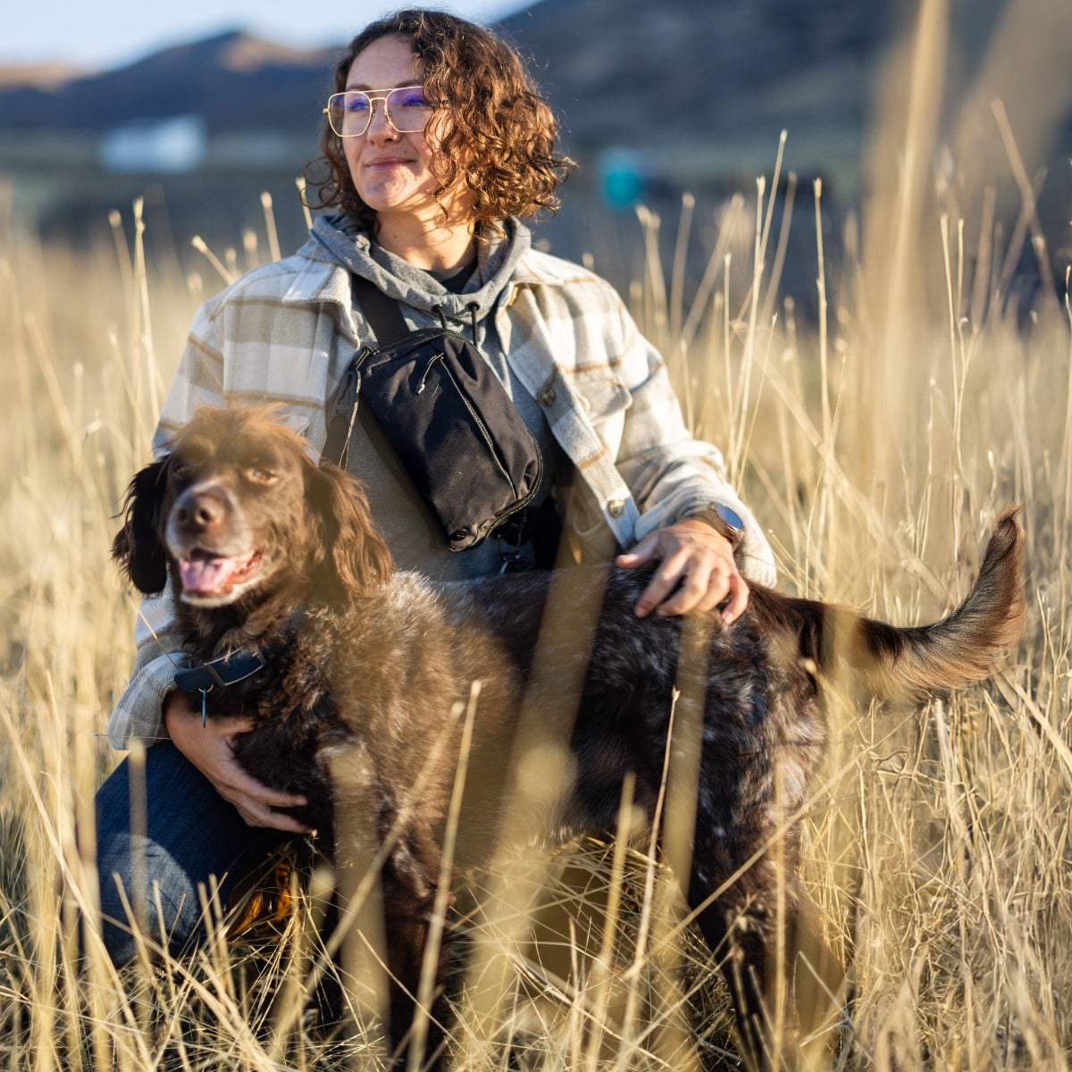 Photograph of a woman with a brown and white dog in a golden field with mountains in the background the woman wears glasses and a plaid shirt the dog has a long tail and is speckled