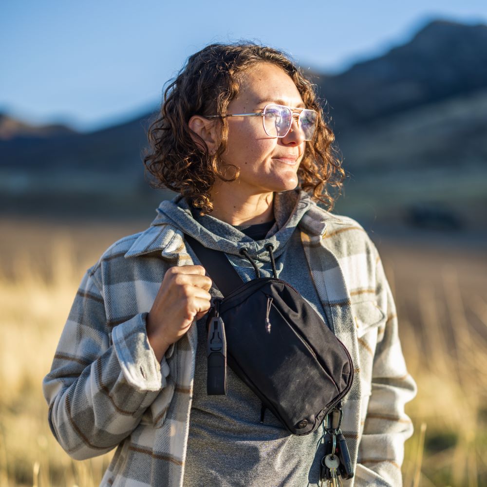 Photograph of a woman in a sun-drenched landscape wearing a grey and white plaid shirt a black crossbody bag and stylish glasses with distant mountains visible