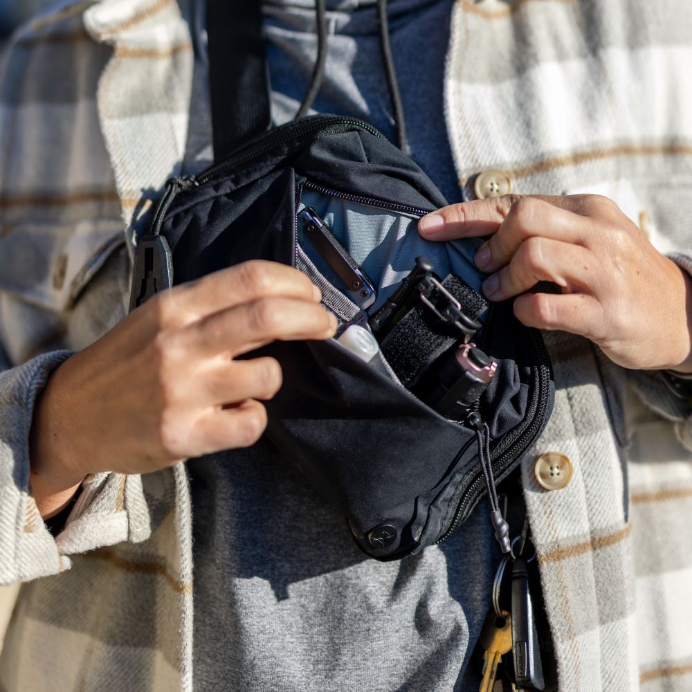 Photograph showing a person's hands unpacking a black sling bag containing a multi-tool, hand sanitizer, and a flashlight with a gray interior and subtle branding details