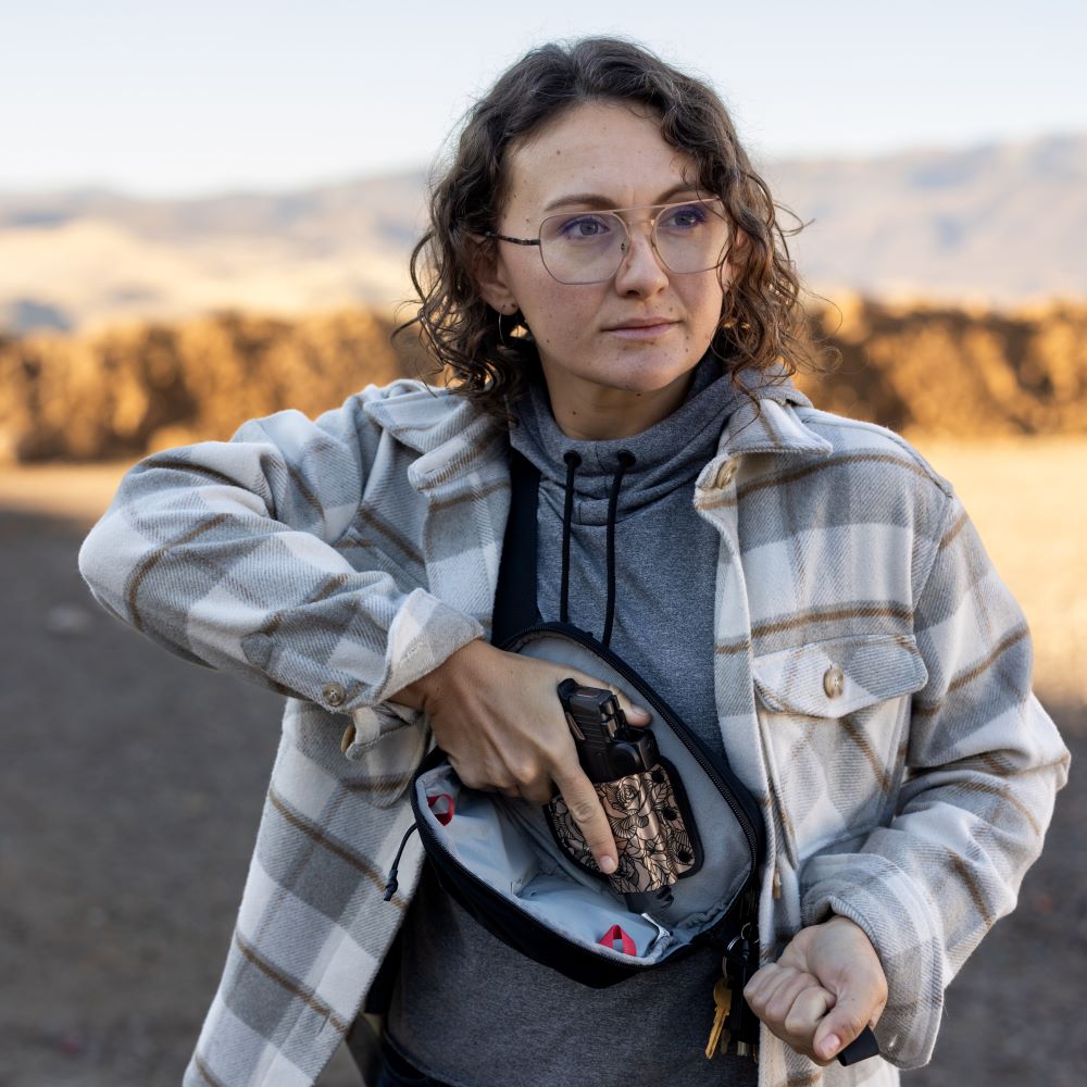 Photograph of a woman drawing a handgun from a floral-print concealed carry purse while wearing glasses and a beige and white plaid shirt against a backdrop of golden hay bales and distant mountains