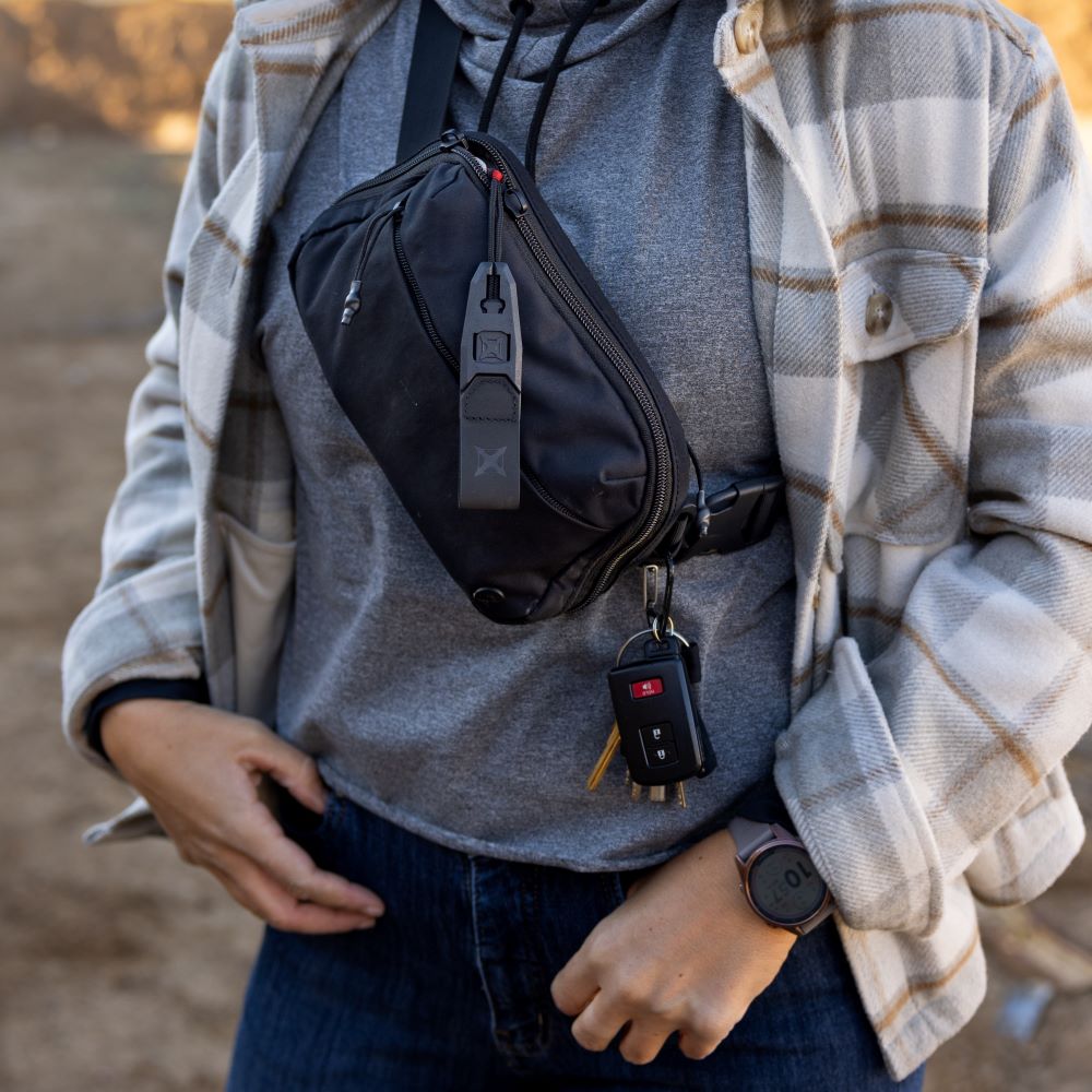 Photograph of a person wearing a black crossbody bag with a gray shirt and beige plaid jacket outdoors showcasing a sleek design, a red accent zipper pull, and a stylish watch.