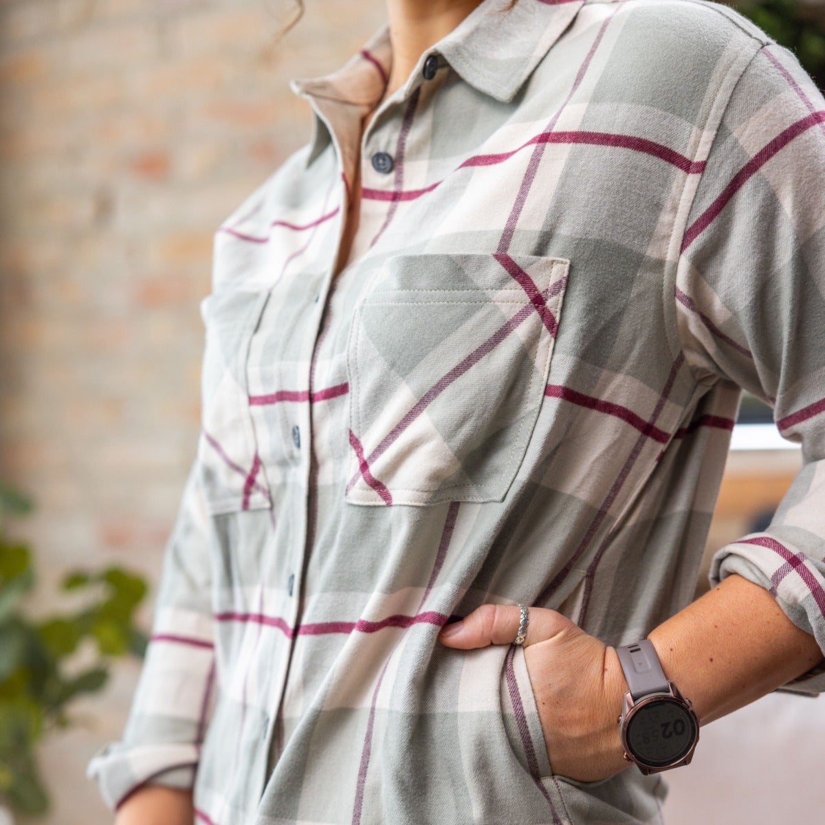 Photograph of a sage green and burgundy plaid shirt with a breast pocket and a woman's hand in the pocket wearing a rose gold watch and ring against a blurred brick wall background