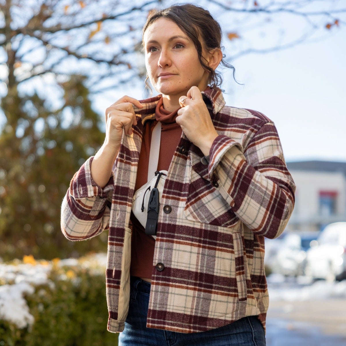 Photograph of a woman wearing a maroon and cream plaid shirt adjusting her collar outdoors near a building and autumn trees with hints of snow and sunlight