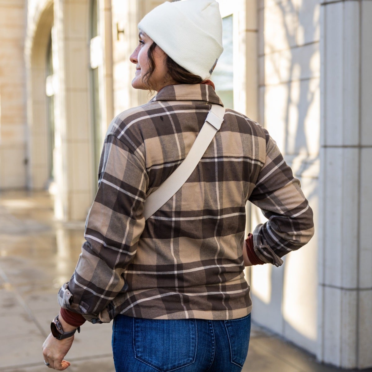 Photograph of a woman wearing a brown and beige plaid shirt, a white beanie, and a cream-colored crossbody bag walking away from the camera in an outdoor urban setting with sunlit building facades and shadows cast on the walls.