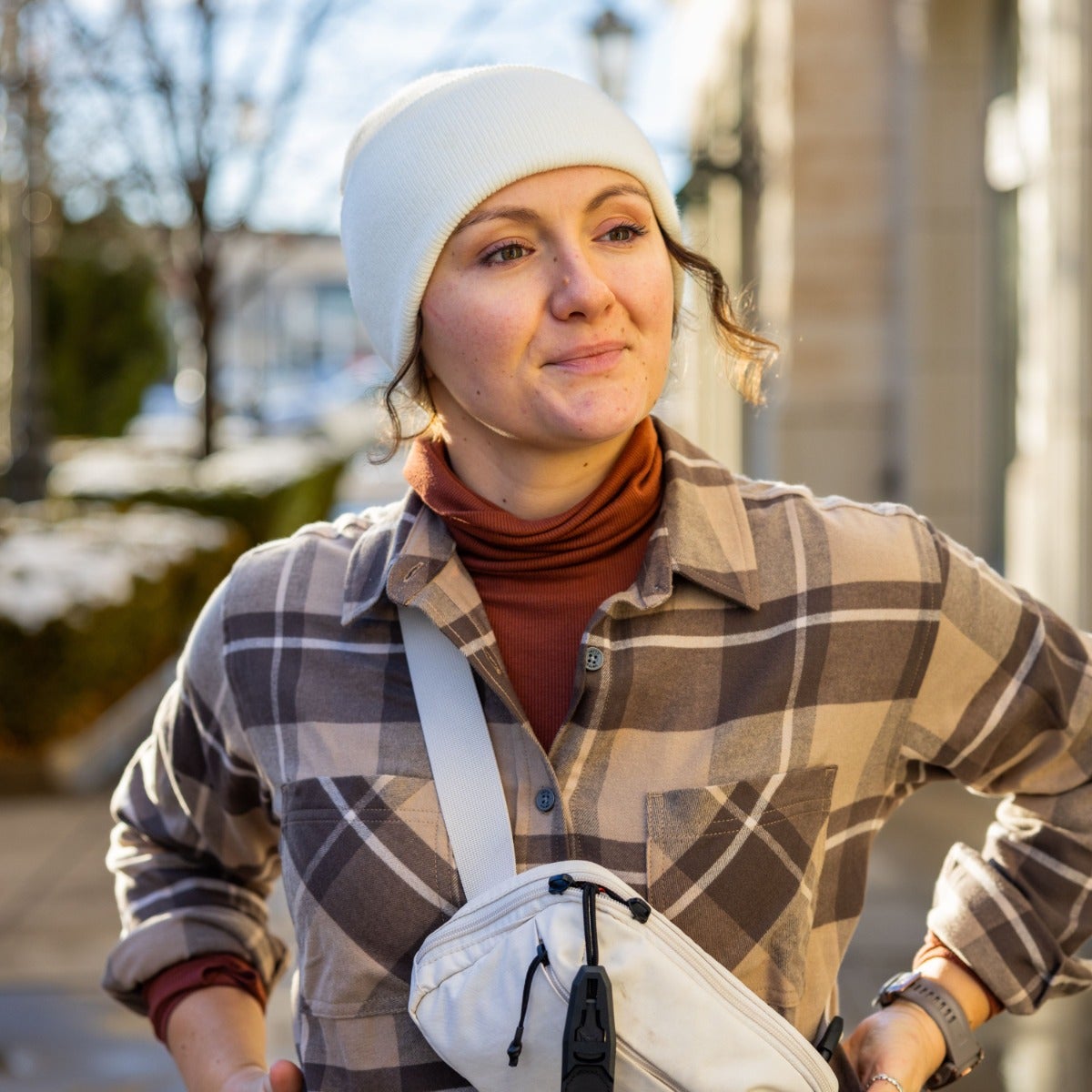 Photograph of a woman wearing a white knit cap, rust turtleneck, and brown and beige plaid shirt with a white crossbody bag on a city street with partially visible buildings and trees in the background